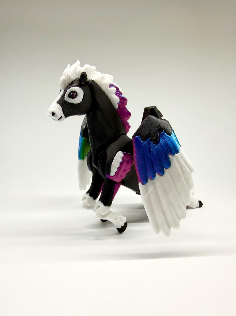May include: A 3D-printed Pegasus figurine. The black horse has white feathered wings with blue and white tips, and a rainbow gradient on the inner wings. The mane and tail are white and purple. The figurine is on a white background.