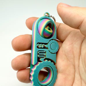 May include: A teal and rainbow-colored keychain with a unique, geometric design. The keychain features a circular gear, a key-like shape, and a spherical element. It is attached to a small metal chain.