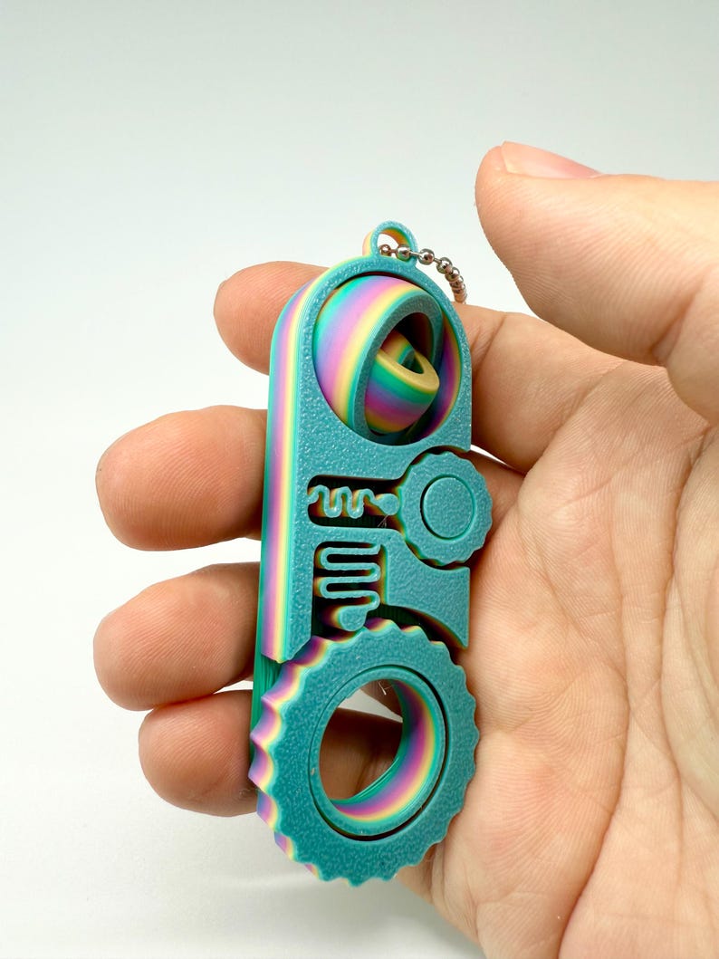 以下が含まれることがあります： ユニークな幾何学模様の、ティールと虹色のキーホルダー。 キーホルダーは、円形の歯車、キーのような形、球状の要素が特徴です。 小さな金属チェーンが付いています。