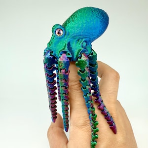 May include: A multi-coloured articulated octopus toy with a metallic finish. The octopus has a blue head, green body, and rainbow-coloured tentacles. The toy is held against a white background, highlighting its vibrant colours and detailed design.
