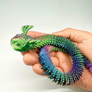 May include: A colorful, articulated dragon toy in shades of green, blue, and purple. The dragon is held in a hand and has a textured, scale-like appearance. The toy's head features small, leaf-like appendages.