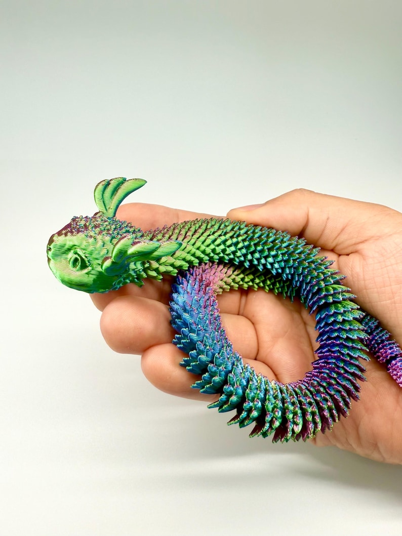 May include: A colorful, articulated dragon toy in shades of green, blue, and purple. The dragon is held in a hand and has a textured, scale-like appearance. The toy's head features small, leaf-like appendages.