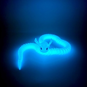 May include: A glowing blue dragon toy with a textured, segmented body and large, expressive eyes. The dragon is curled in a relaxed pose against a dark background, emitting a soft, ethereal light. The toy's design suggests a fantasy or mythical theme.
