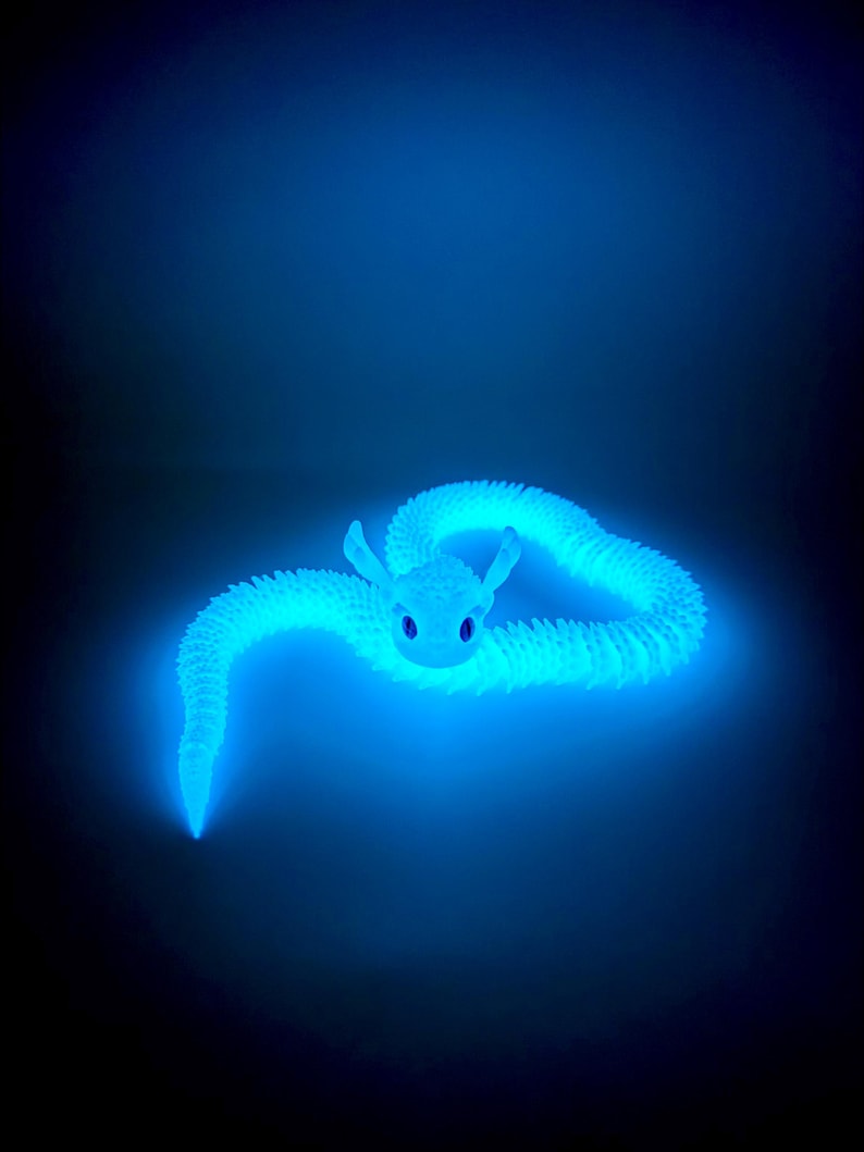 May include: A glowing blue dragon toy with a textured, segmented body and large, expressive eyes. The dragon is curled in a relaxed pose against a dark background, emitting a soft, ethereal light. The toy's design suggests a fantasy or mythical theme.