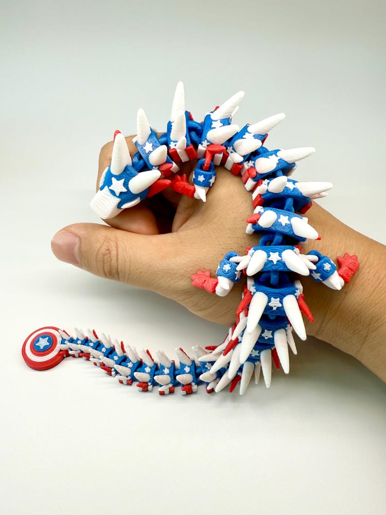 Puede incluir: Un juguete de drag&oacute;n articulado y flexible en rojo, blanco y azul. El cuerpo del drag&oacute;n est&aacute; segmentado, con cada segmento con detalles de estrellas blancas sobre un fondo azul. La cola del drag&oacute;n tiene un dise&ntilde;o de escudo rojo y blanco.