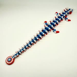 Puede incluir: Un juguete articulado largo, rojo, blanco y azul, que se asemeja a una columna vertebral o a una criatura segmentada. El juguete presenta un escudo del Capit&aacute;n Am&eacute;rica en un extremo. Los segmentos son azules con estrellas blancas y detalles rojos.
