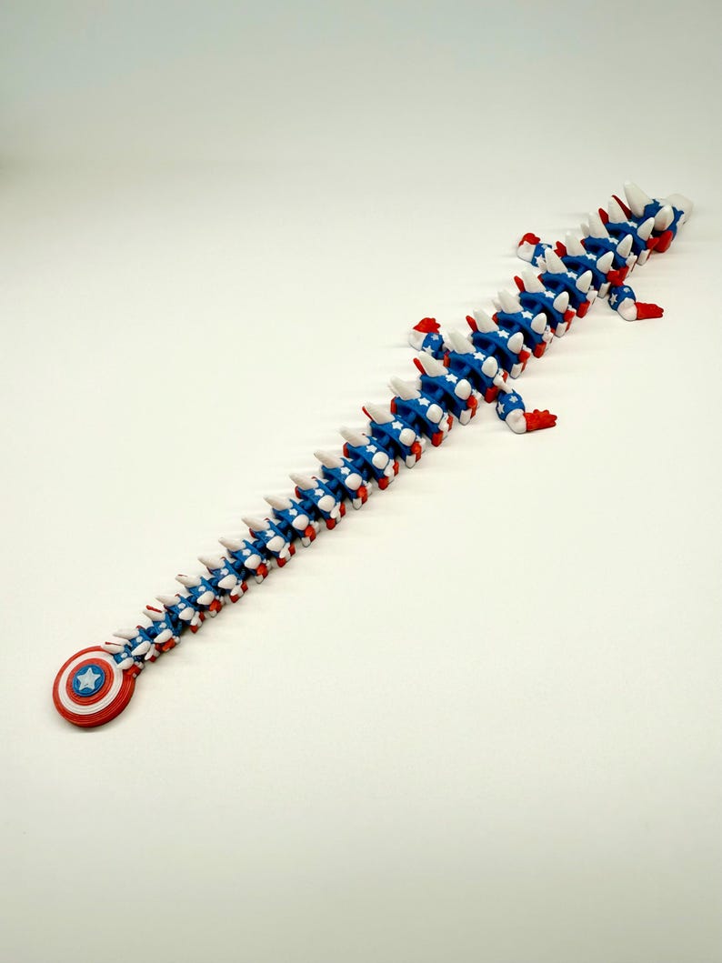 Puede incluir: Un juguete articulado largo, rojo, blanco y azul, que se asemeja a una columna vertebral o a una criatura segmentada. El juguete presenta un escudo del Capit&aacute;n Am&eacute;rica en un extremo. Los segmentos son azules con estrellas blancas y detalles rojos.