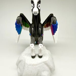 May include: A black and white pegasus figurine with outstretched wings. The wings have white feathers with colorful accents of blue, green, and purple. The figurine stands on a gray, textured rock-like base.