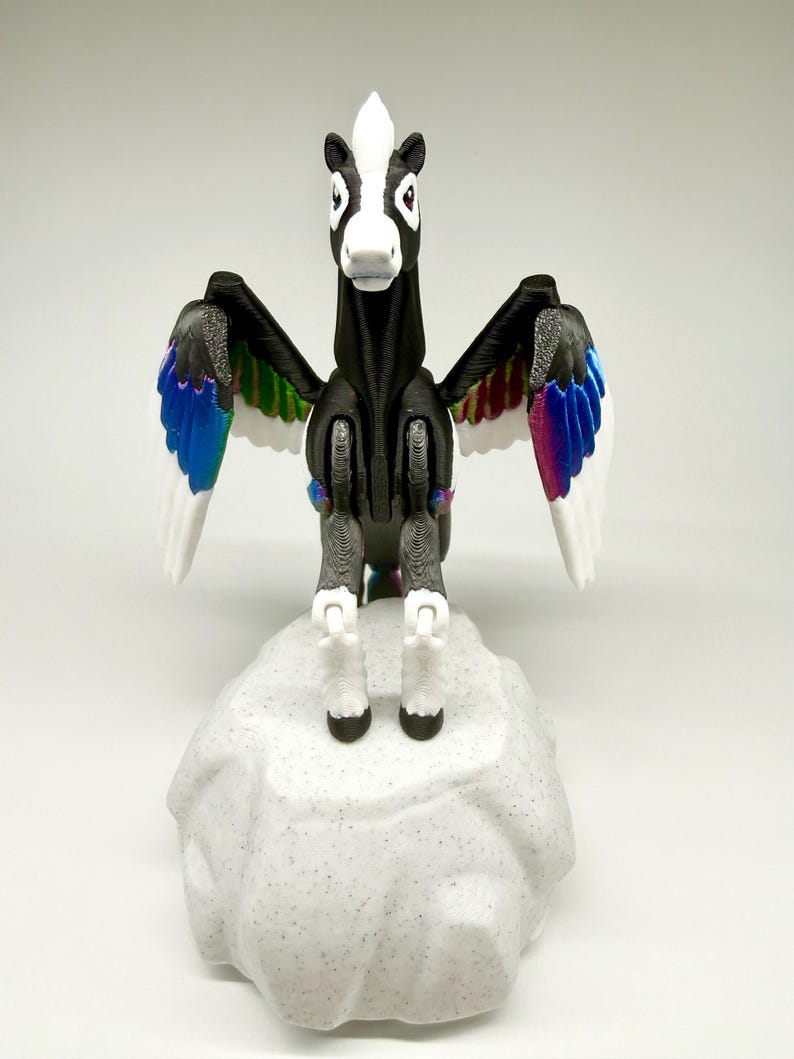May include: A black and white pegasus figurine with outstretched wings. The wings have white feathers with colorful accents of blue, green, and purple. The figurine stands on a gray, textured rock-like base.