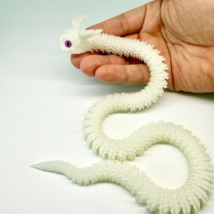 May include: A white, articulated dragon toy with a textured, scale-like body. The dragon has a curved shape, with a head featuring small horns and purple eyes. The toy is held in a hand against a white background.
