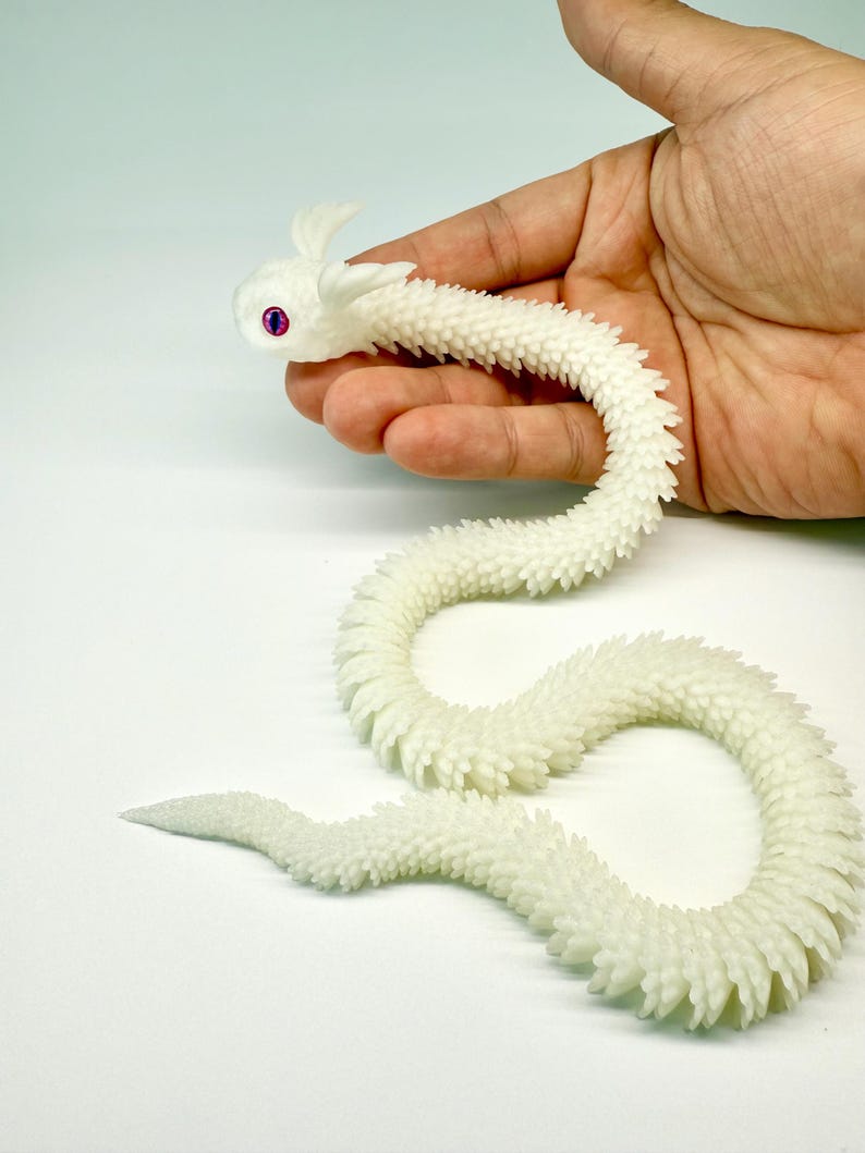 May include: A white, articulated dragon toy with a textured, scale-like body. The dragon has a curved shape, with a head featuring small horns and purple eyes. The toy is held in a hand against a white background.