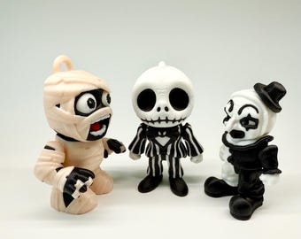 May include: Three Halloween-themed figurines: a mummy, a skeleton in a pinstripe suit, and a clown. The mummy is tan with black accents, the skeleton is white and black, and the clown is black and white. All three have exaggerated features.