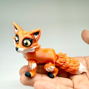 Puede incluir: Una figura de zorro naranja impresa en 3D con pecho y punta de la cola blancos, patas negras y ojos grandes. El zorro tiene articulaciones articuladas y un diseño detallado y texturizado. La figura se sostiene en una mano.