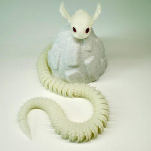 May include: A white, fantasy-themed sculpture of a creature with a snake-like body and a head with large ears and red eyes. The creature is perched on a textured, light gray rock. The snake-like body is segmented and curves across a white surface.