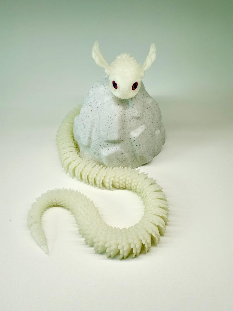May include: A white, fantasy-themed sculpture of a creature with a snake-like body and a head with large ears and red eyes. The creature is perched on a textured, light gray rock. The snake-like body is segmented and curves across a white surface.