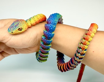 Op de afbeelding: Een kleurrijk, beweeglijk slangen speelgoed met een regenboogverloop. De kop is geel en groen, overgaand in blauw, rood en oranje langs het lichaam. Het speelgoed is om een pols gewikkeld.