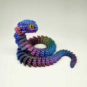 May include: A colorful, iridescent snake sculpture. The snake is primarily blue, purple, and green, with a textured, scale-like pattern. The head features a large, detailed eye with yellow and orange accents. The sculpture is posed in a coiled position against a white background.
