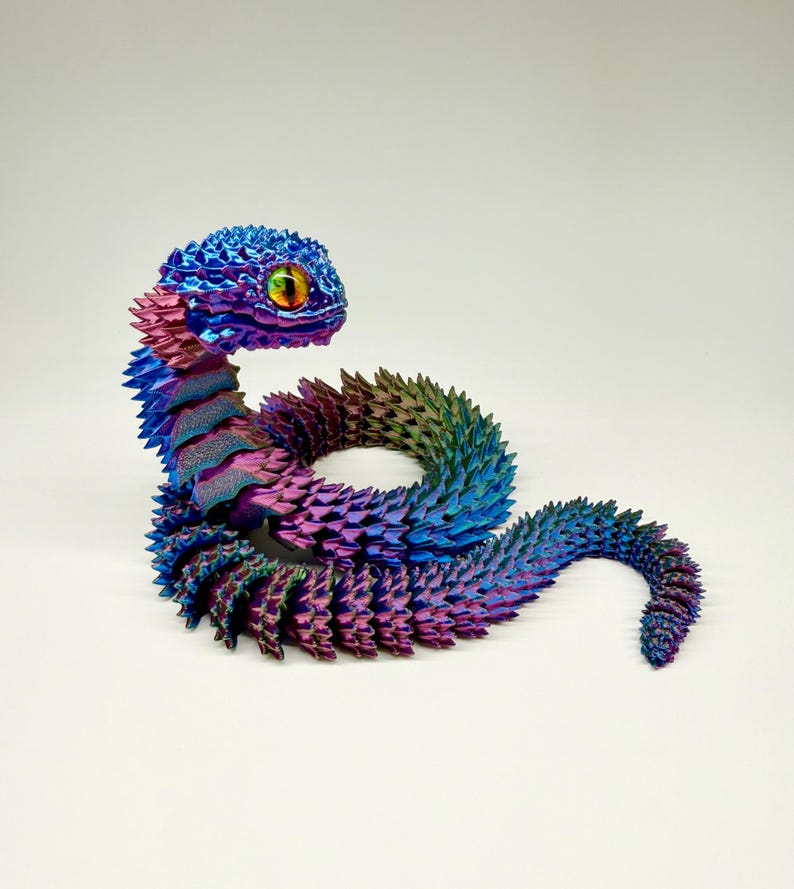 May include: A colorful, iridescent snake sculpture. The snake is primarily blue, purple, and green, with a textured, scale-like pattern. The head features a large, detailed eye with yellow and orange accents. The sculpture is posed in a coiled position against a white background.