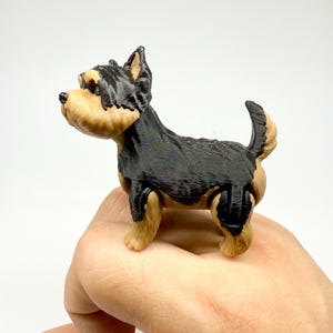 May include: A small, 3D-printed Yorkshire Terrier figurine. The dog has a black body, tan legs, and a tan and black head. The figurine is held in a hand against a white background.