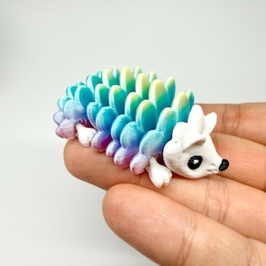 May include: A 3D-printed articulated hedgehog toy. The body is a gradient of blue, green, and purple. The head and feet are white with black eyes and a black nose. The toy is held in a hand.