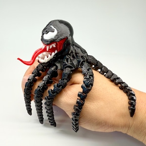 Könnte beinhalten: Eine schwarze, bewegliche Venom-Figur mit roter Zunge und weißen Zähnen. Die Figur hat mehrere gegliederte Beine und einen weißen Fleck auf dem Kopf. Die Figur wird in einer Hand vor weißem Hintergrund gehalten.