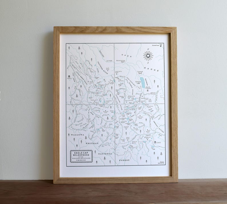May include: A framed map print of the Eagle Cap Wilderness in the Wallowa Mountains. The map is in shades of blue and white, with detailed geographical features and text labels. The frame is made of light-colored wood.