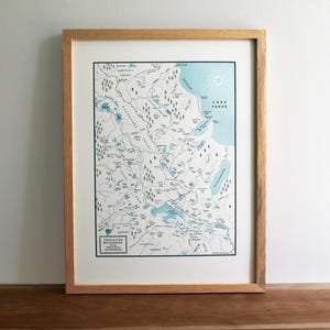 May include: Framed map print of the Desolation Wilderness in California, featuring a detailed illustration in shades of blue and white. The map is encased in a light brown wooden frame, showcasing the topography and landmarks of the area.
