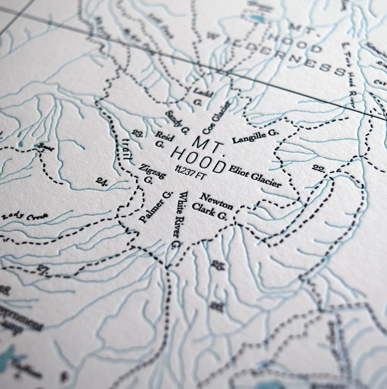 Puede incluir: Un primer plano de un mapa que muestra la ubicaci&oacute;n del Monte Hood, Oreg&oacute;n. El mapa est&aacute; impreso en blanco y negro sobre un fondo blanco. La elevaci&oacute;n del Monte Hood se indica como 11.237 pies. El mapa muestra la ubicaci&oacute;n de varios glaciares, incluidos el glaciar Coe, el glaciar Eliot y el glaciar Langille.