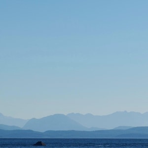 May include: A tranquil seascape with a wide expanse of blue water beneath a clear, pale blue sky. Distant mountains recede into the horizon, creating depth. A small, dark rock formation is in the water.
