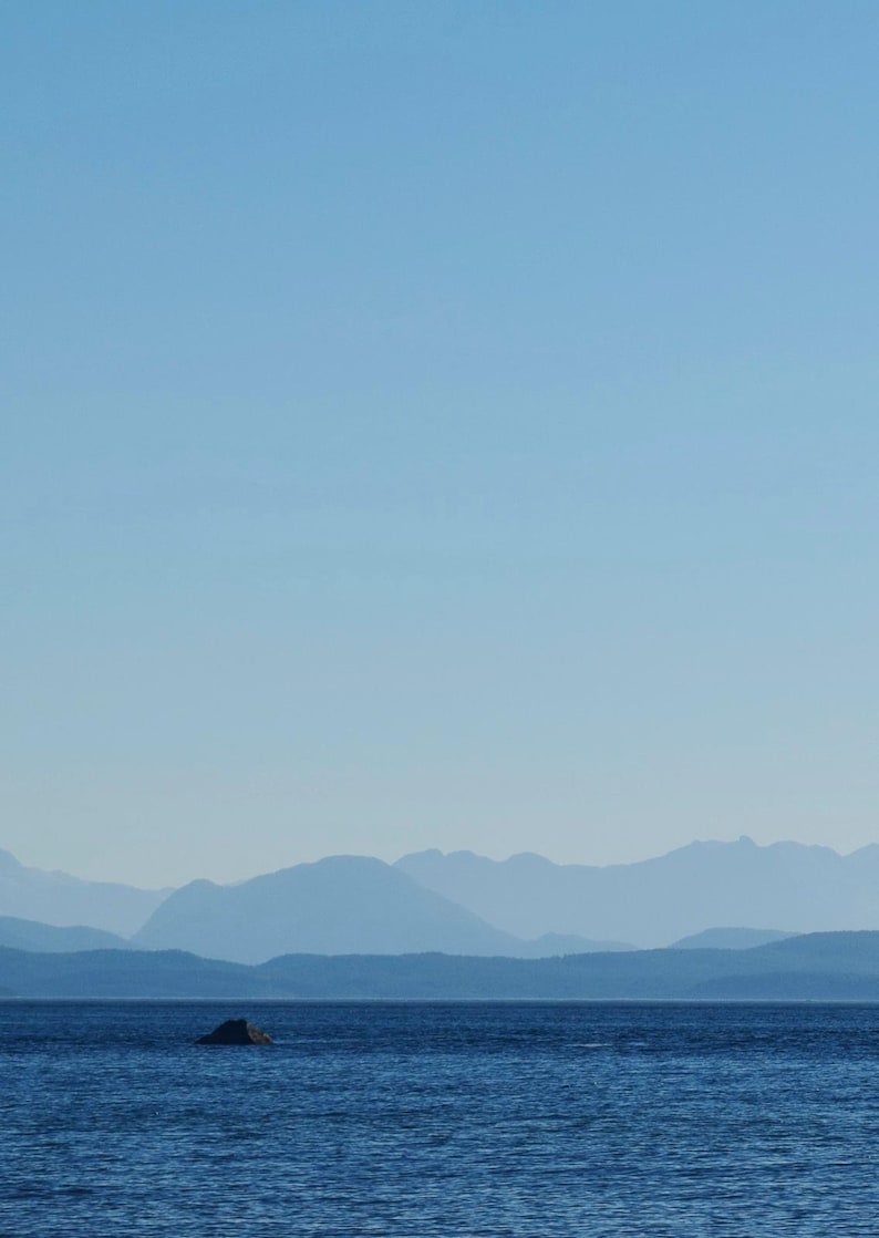 May include: A tranquil seascape with a wide expanse of blue water beneath a clear, pale blue sky. Distant mountains recede into the horizon, creating depth. A small, dark rock formation is in the water.