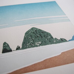 May include: A print of a coastal landscape featuring a large, textured green rock formation in the center, with smaller rocks to the left. The sky is light blue with white streaks, and the foreground shows a sandy beach.