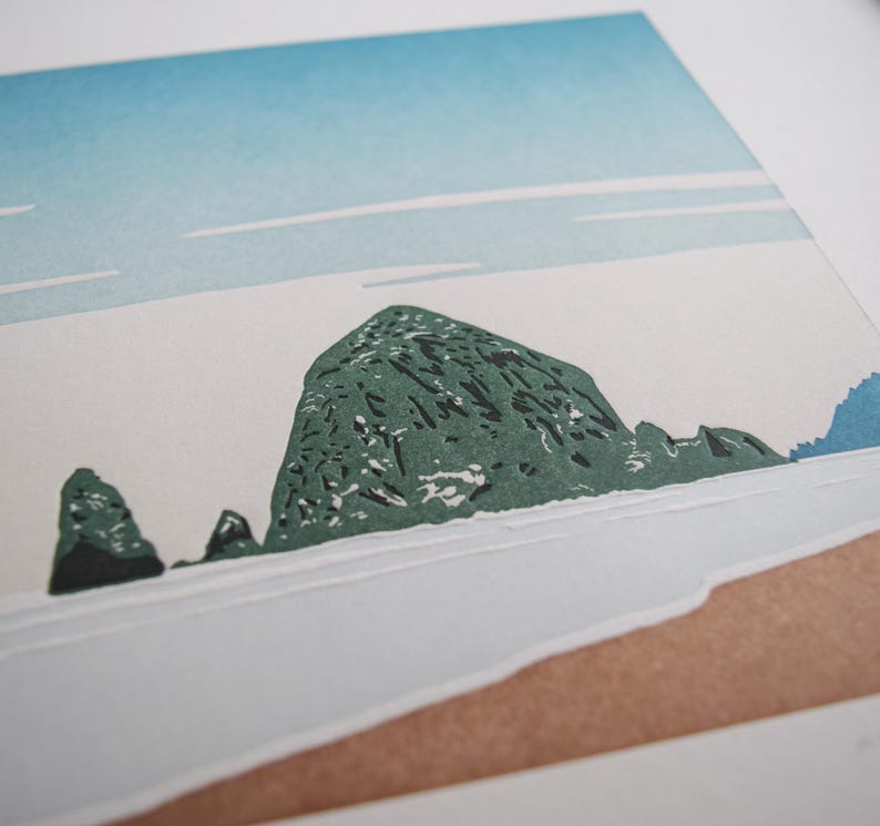 May include: A print of a coastal landscape featuring a large, textured green rock formation in the center, with smaller rocks to the left. The sky is light blue with white streaks, and the foreground shows a sandy beach.