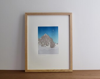 May include: Framed art print depicting a mountain landscape under a blue sky with a crescent moon. The artwork is in a wooden frame and has a white border. The mountain is rendered in shades of grey and white.