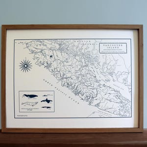 May include: A framed map print of Vancouver Island, British Columbia, with detailed geographical outlines and text. The map is in black and white, with a compass rose and whale illustrations. The frame is made of wood.