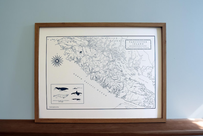 May include: A framed map print of Vancouver Island, British Columbia, with detailed geographical outlines and text. The map is in black and white, with a compass rose and whale illustrations. The frame is made of wood.