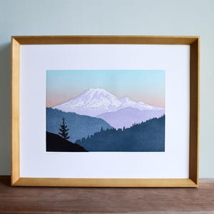 May include: A framed art print featuring a mountain landscape. The print showcases a snow-capped mountain peak, with layers of blue and purple mountains in the background. The sky transitions from light blue to pink. The frame is a light brown color.