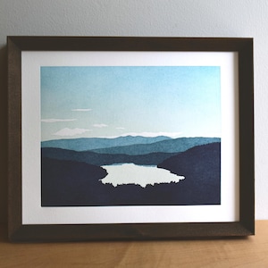 May include: A framed landscape print featuring a layered mountain range in shades of dark blue and navy, overlooking a central lake with a light-colored surface. The sky is a pale blue with thin white clouds, all enclosed in a dark brown frame.