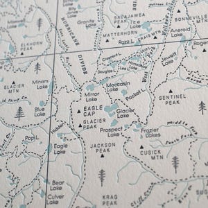 May include: Close-up of a detailed map with topographical features. The map features names of lakes, mountains, and rivers in a light blue color, with black dashed lines indicating trails and borders. The map is printed on a textured, off-white paper.