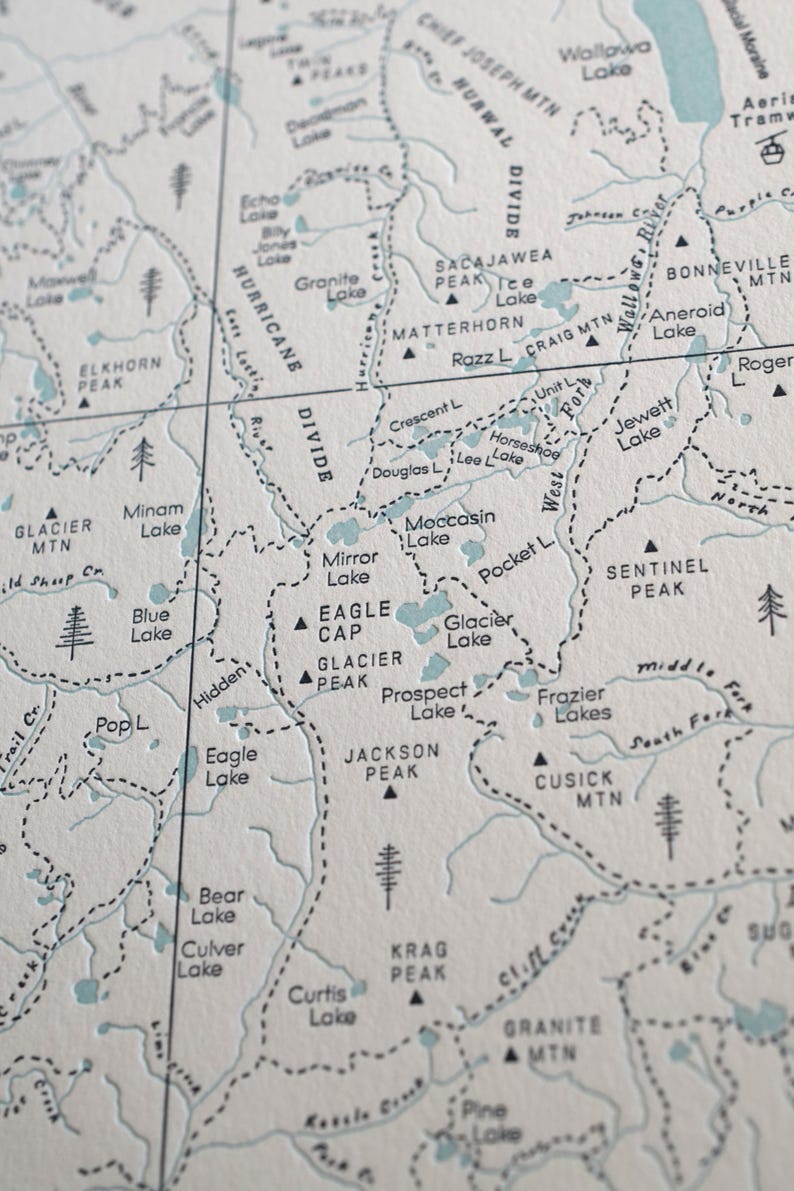 May include: Close-up of a detailed map with topographical features. The map features names of lakes, mountains, and rivers in a light blue color, with black dashed lines indicating trails and borders. The map is printed on a textured, off-white paper.