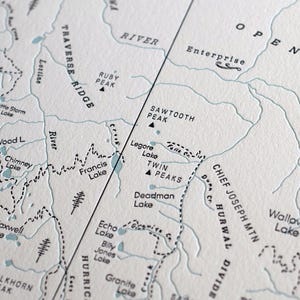 May include: Close-up of a detailed map with topographical features. The map features names like "Sawtooth Peak," "Francis Lake," and "Enterprise." The color palette is muted, with a cream background and blue-gray lines, creating a vintage aesthetic.