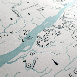 May include: Detailed map print featuring the Columbia River, with Washington and Oregon labeled. The map includes hiking trails, landmarks, and the names Cape Horn, Angels Rest, and Devils Rest. The color palette is primarily white and light blue.