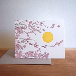 May include: A white greeting card featuring a yellow sun and pink cherry blossom branches. The card rests on a brown surface with a grey envelope. The design is a minimalist nature illustration.
