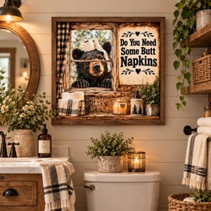 May include: A square wall art piece in a wooden frame featuring a black bear looking through a window with a roll of toilet paper on its head. The text reads Do You Need Some Butt Napkins. The image includes a wire basket with toilet paper.