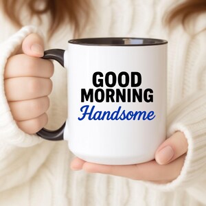 May include: White ceramic mug with a black handle and interior. The mug features the text "GOOD MORNING Handsome" in black and blue lettering. The mug is held by a person wearing a white sweater.