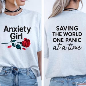 May include: A light gray short-sleeve t-shirt featuring the black text Anxiety Girl and a red rose on the front. The back of the shirt displays the black text SAVING THE WORLD ONE PANIC at a time in a combination of bold and script fonts.