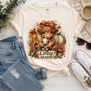 May include: A cream-colored t-shirt featuring a graphic of a fluffy brown Highland cow holding a smaller calf, surrounded by pumpkins, sunflowers, and autumn foliage. The text "Cozy Season" is printed on a wooden sign below the animals.