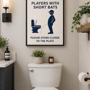 May include: A framed bathroom art print with the text "PLAYERS WITH SHORT BATS PLEASE STAND CLOSER TO THE PLATE." The artwork features a navy blue silhouette of a person urinating into a toilet. The print is displayed above a white toilet.