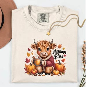 May include: Cream-colored Comfort Colors XL t-shirt featuring a cartoon Highland cow wearing a sweater and holding a mug. The design includes pumpkins, fall leaves, and the text "Autumn Bliss". A gold star pendant necklace is draped across the shirt.