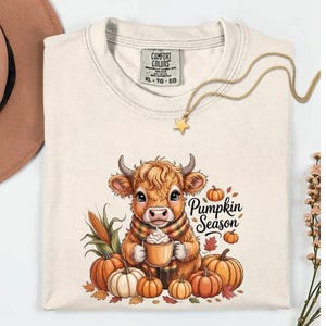 May include: Cream-colored t-shirt with a graphic of a Highland cow wearing a scarf and holding a mug. The design includes pumpkins, leaves, and the text "Pumpkin Season". A gold necklace with a star pendant is also visible.