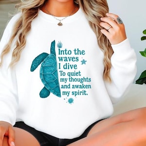May include: White sweatshirt featuring a teal turtle graphic and the text "Into the waves I dive To quiet my thoughts and awaken my spirit." The turtle design is detailed with textured shading.